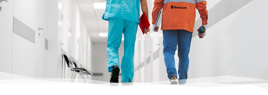 Two individuals walking down a healthcare facility hallway, one in blue scrubs holding a clipboard, one in construction clothing with Terracon on the back of the safety vest