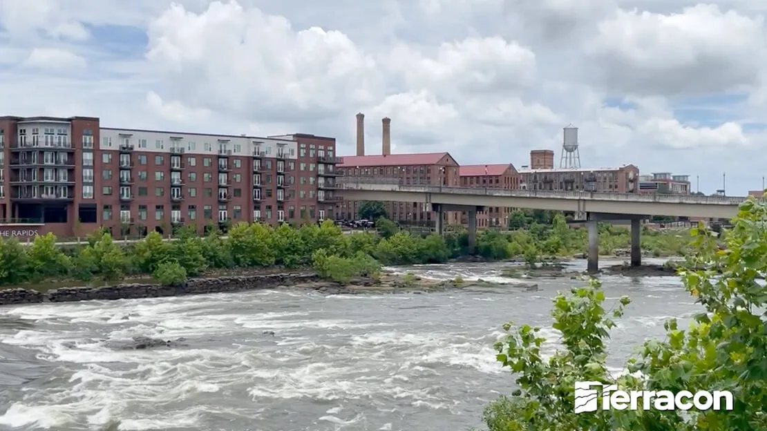 River rapids flowing beneath concrete bridge with brick mill buildings, apartments, and water tower in background