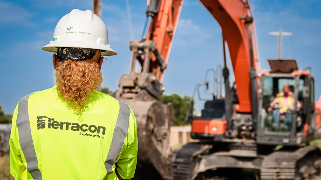 Terracon employee with red hair wearing a hi-vis vest and a hard hat with her back to the camera, looking at a backhoe.